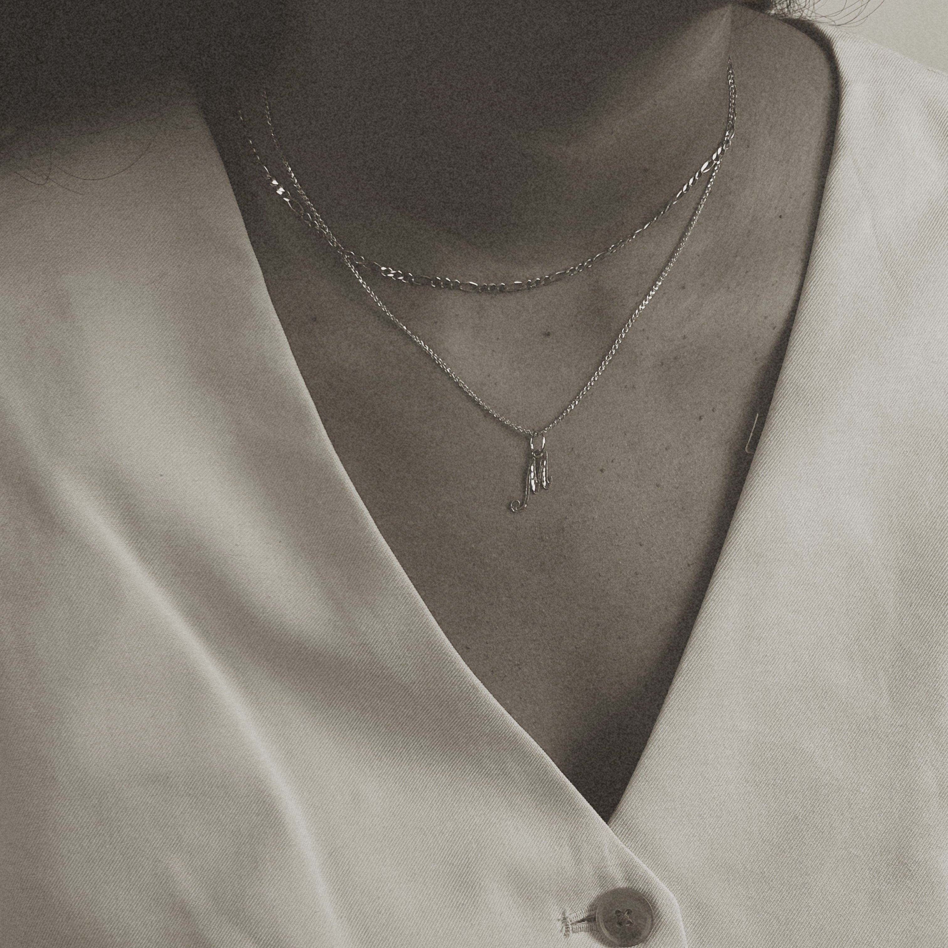 image of a women with two nechlaces. one chain is a gold figaro chain and the second is a gold letter chain with the pendant m