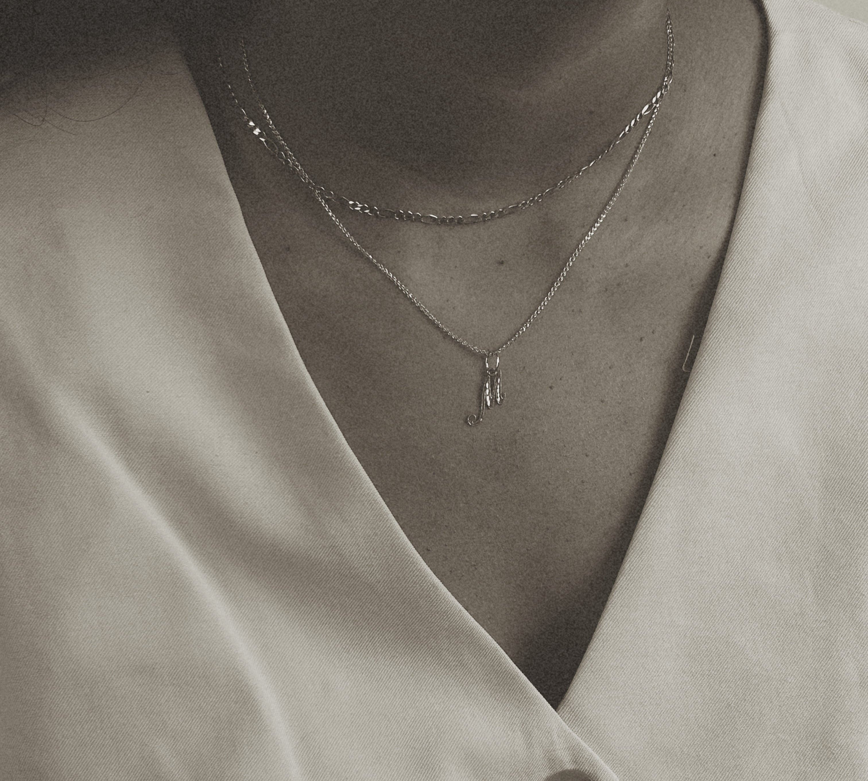 image of a women with two nechlaces. one chain is a gold figaro chain and the second is a gold letter chain with the pendant m