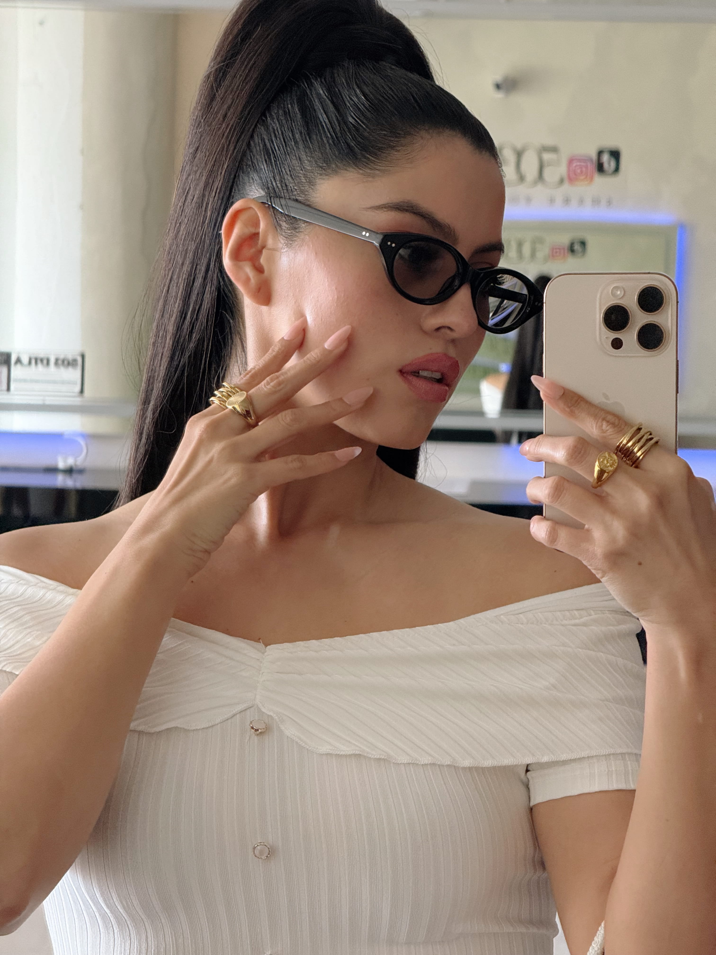 Woman taking a mirror selfie wearing sunglasses and a white off-shoulder top.