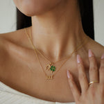 Close-up of a person wearing a gold necklace with a clover pendant and a ring on a neutral background