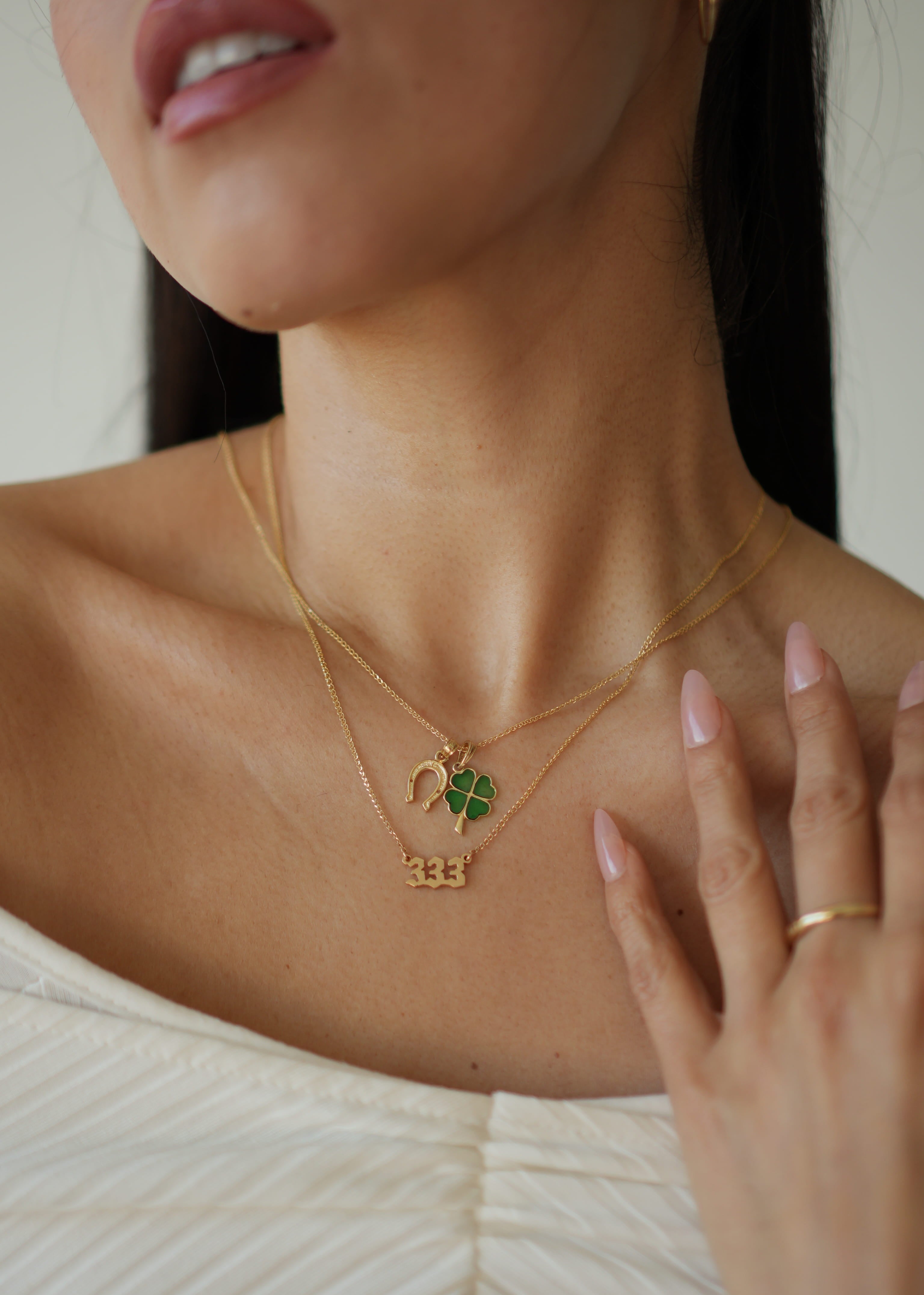 Close-up of a person wearing a gold necklace with a clover pendant and a ring on a neutral background