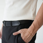 Person wearing a white shirt, black pants, and a black belt with a gold bracelet on a plain background