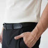 Person wearing a white shirt, black pants, and a black belt with a gold bracelet on a plain background