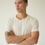 Man wearing a white t-shirt and black pants with arms crossed against a plain background
