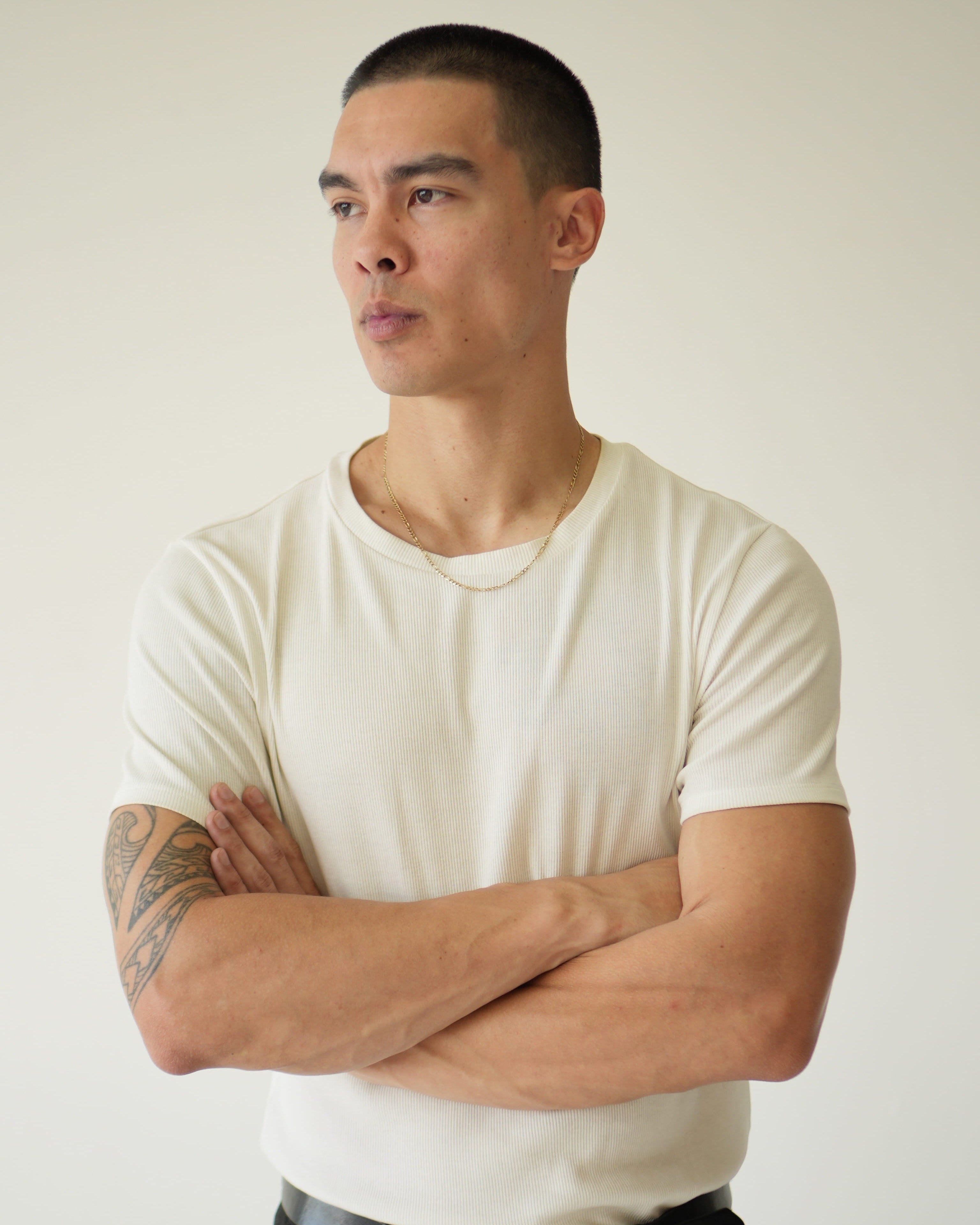 Man wearing a white t-shirt and black pants with arms crossed against a plain background