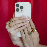 Hand holding a white smartphone with gold rings on fingers against a red background