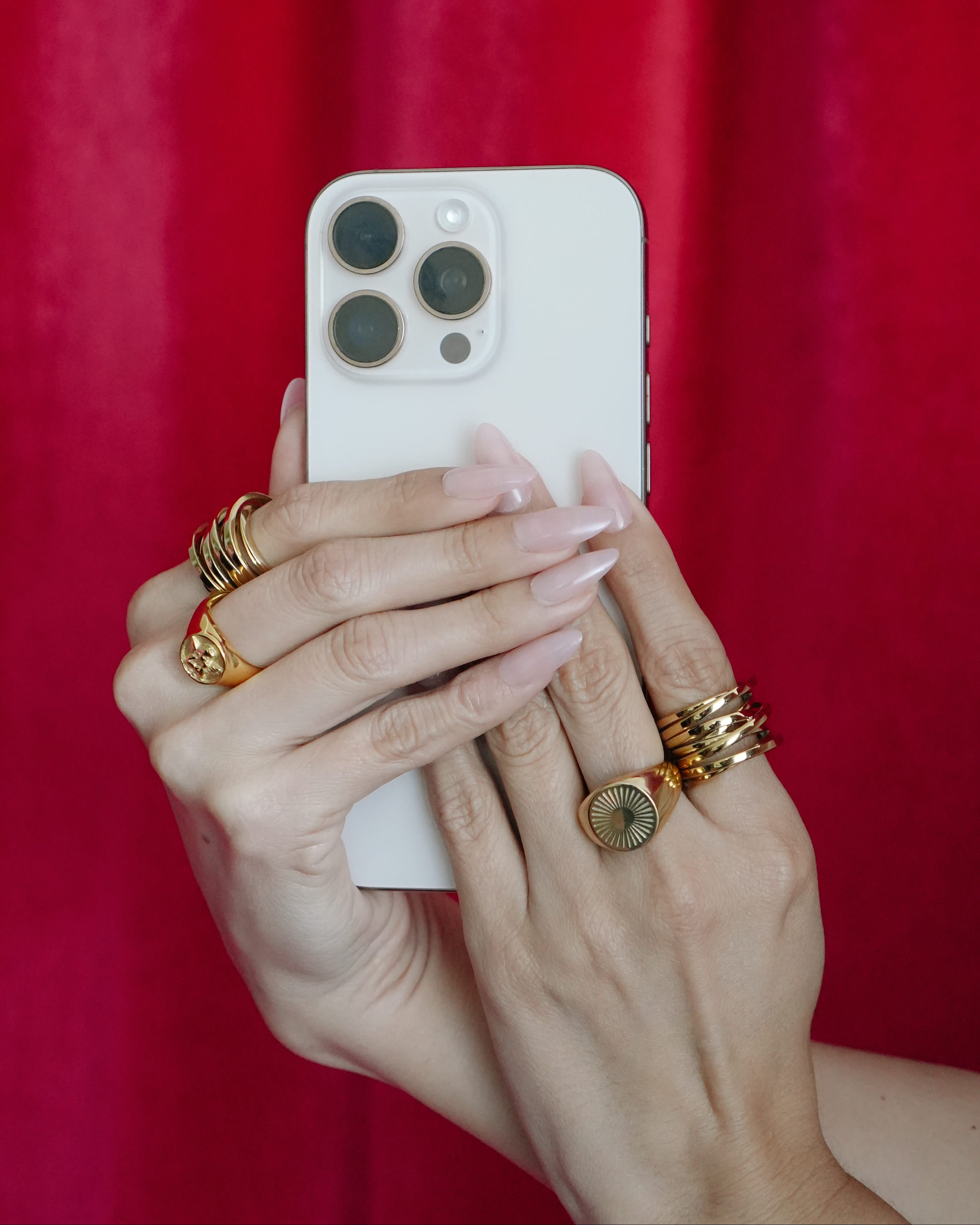 Hand holding a white smartphone with gold rings on fingers against a red background